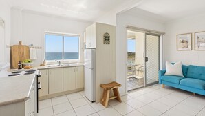 Una cocina luminosa con vistas al mar, un taburete de madera, un sofá azul y una zona de comedor con mesa y sillas.