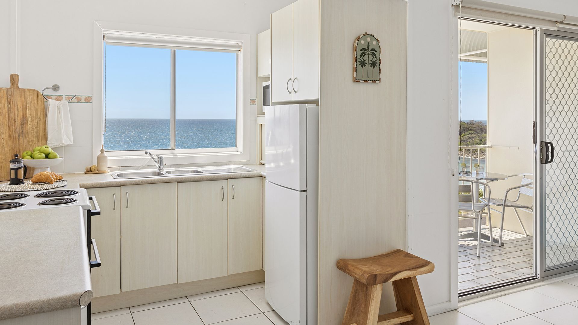 A kitchen with a view of the sea, a refrigerator, a wooden stool, and a dining area with a table and chairs.