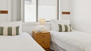 A hotel room with two beds, a wooden nightstand with a lamp, and a window with a metal screen.
