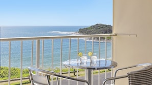 A balcony with a table and chairs overlooking a beach with clear blue water and a distant rocky island.