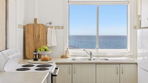 Una cocina con vista al océano, una tabla de cortar de madera, un bol de manzanas verdes, una cafetera y un croissant en la encimera.