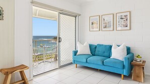Habitación con sofá azul, taburete de madera, puerta corrediza de vidrio que da a un balcón con vista al océano y tres cuadros en la pared.