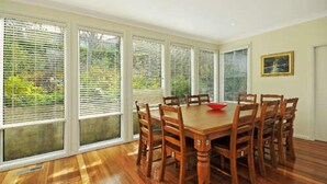 A dining room with a wooden table and chairs, large windows with blinds, and a framed picture on the wall. - Tranquility Large Mountain Home With Views (KATOOMBA)