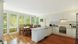 A modern kitchen with white cabinets, a wooden dining table, and large windows. - Tranquility Large Mountain Home With Views (KATOOMBA)