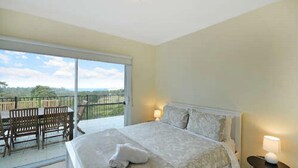 A bedroom with a large bed, a balcony with a table and chairs, and a view of the landscape. - Tranquility Large Mountain Home With Views (KATOOMBA)