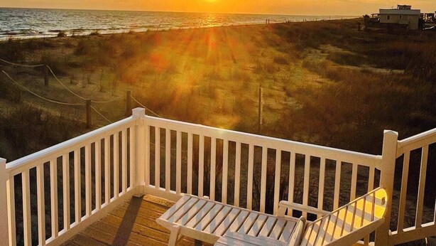 A deck with two chairs and a table, overlooking a beach and ocean during sunset.