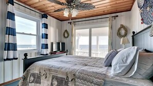 A bedroom with a large bed, a ceiling fan, a TV, and a view of the ocean.