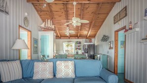 A cozy room with a blue sofa, wooden ceiling beams, a ceiling fan, and a kitchen area with a refrigerator.