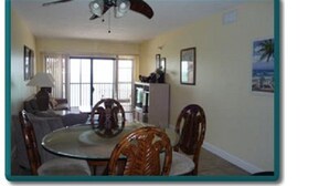 A dining area with a round table and chairs, a ceiling fan, and a view of the outdoors through large windows.
