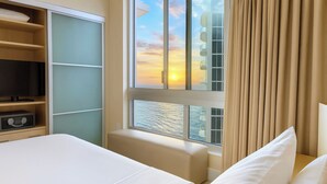 A hotel room with a bed, a seating area, a television, and a window with a view of the sunset. - Oceanfront Condominium with Breathtaking Views and Beach Access (Sunny Isles Beach)