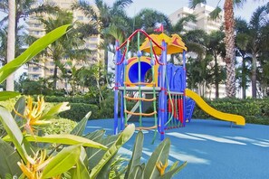 Un terrain de jeux avec des toboggans et des structures d’escalade, entouré de palmiers et d’une zone de jeux bleue.