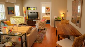A cozy living room with a sofa, armchair, and a dining table. A television is mounted on the wall, and there are paintings on the walls.