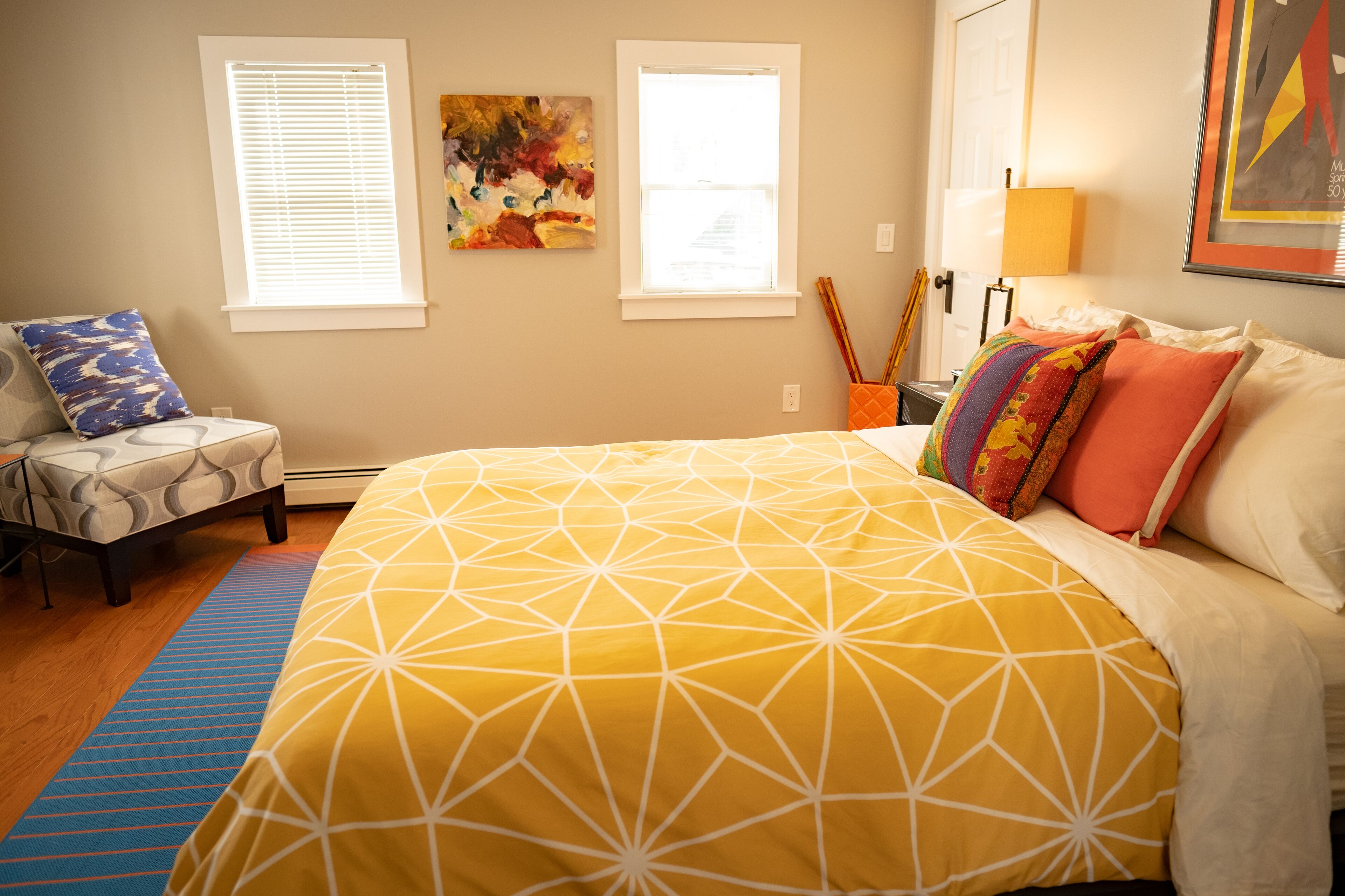 A bedroom with a large bed, two windows, a painting, and a lamp.