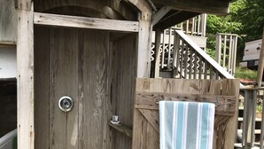 Eine hölzerne Außendusche mit einer Metalttür, ein Handtuch an einer Holzkonstruktion und eine Treppe, die zu einer Terrasse führt.