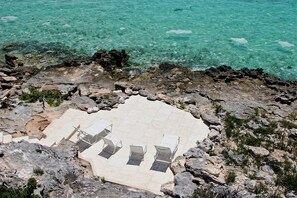 A rocky beach with a tiled area featuring four lounge chairs and a metal railing.