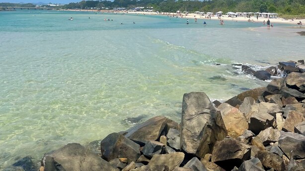Una playa con agua turquesa cristalina, primer plano rocoso y una línea de costa distante con gente en la playa.