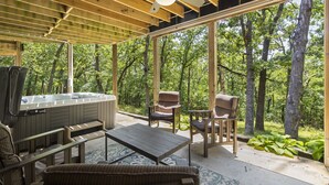 A covered outdoor area with a hot tub, seating, and a view of the forest.