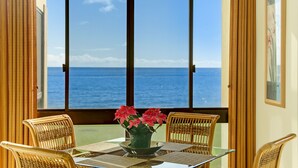 A dining area with a table and chairs, a vase with flowers, and a view of the ocean through large windows. - Poipu Oceanfront, 10 steps to ocean (Poipu)