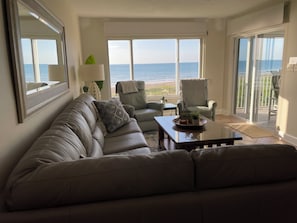 Un salon comprenant un canapé, des fauteuils, une table basse et offrant une vue sur l’océan grâce à de grandes fenêtres.