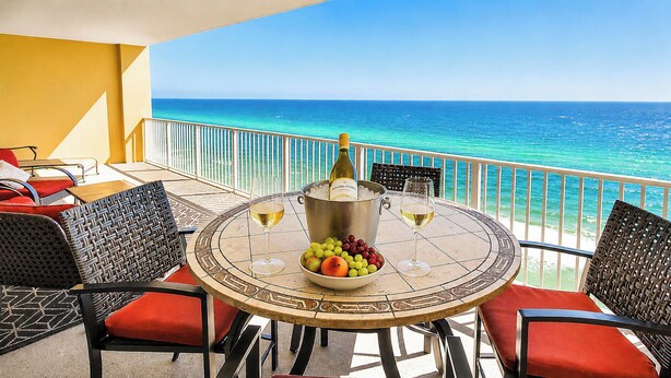 A round table with a bottle of wine, glasses, and a bowl of fruit on a balcony overlooking the ocean.