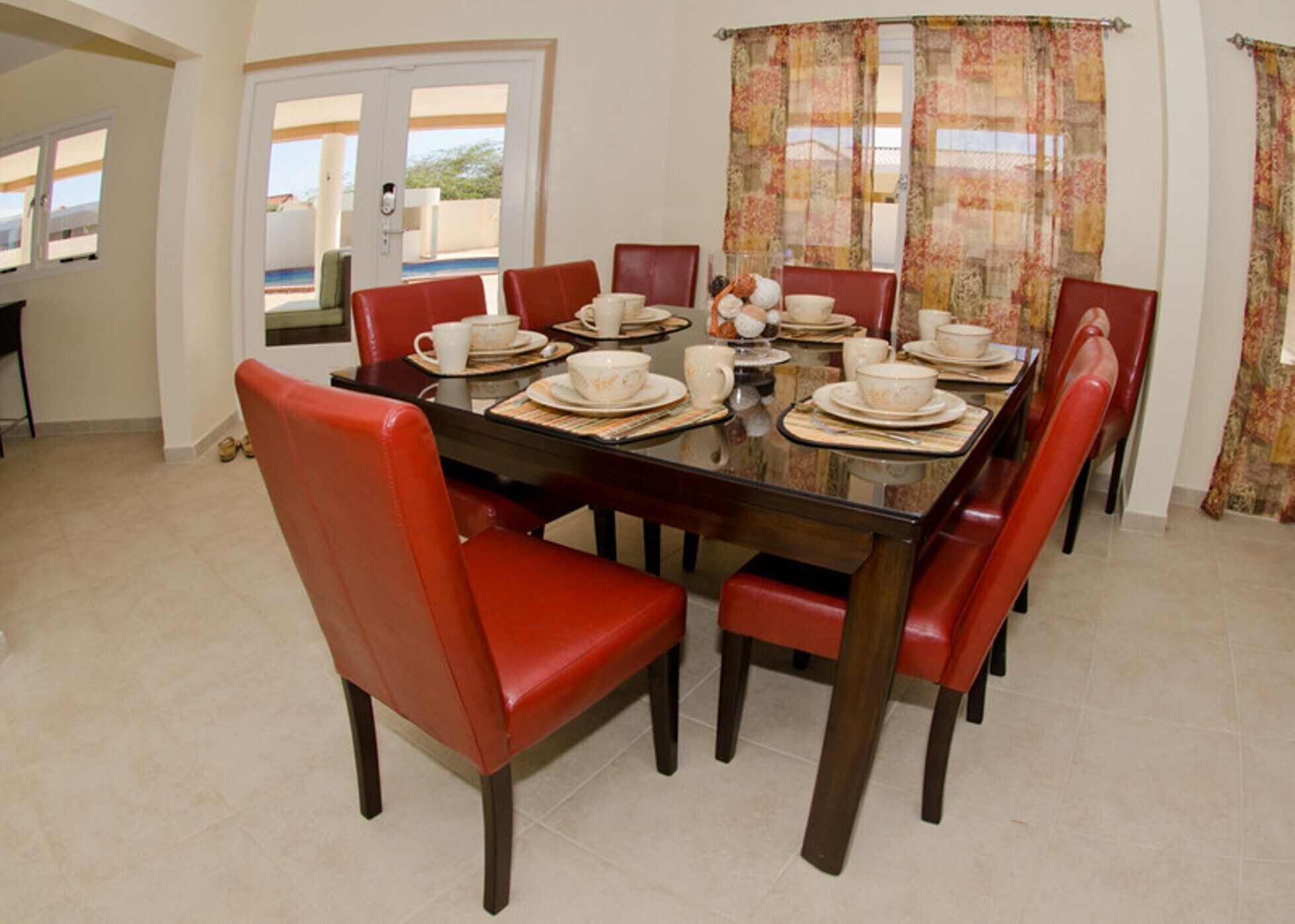Un coin repas avec une table en verre, des chaises rouges et une vue sur l’extérieur par une porte.
