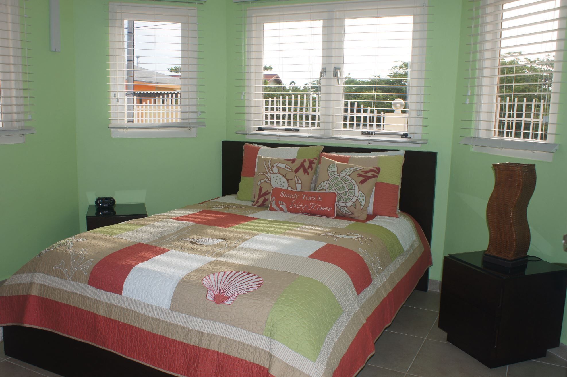 A bedroom with a bed, a nightstand, a lamp, and a window with a view of a white fence and trees.