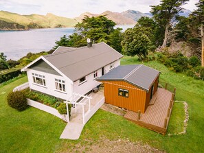 Una casa de dos pisos con un amplio balcón y una estructura de madera separada, rodeada de vegetación exuberante y una vista panorámica de un lago y montañas.