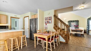 A spacious kitchen with a central island, wooden stools, a dining table with chairs, a refrigerator, and a staircase leading to another level.