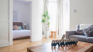 Ein modernes Hotelzimmer mit Bett, Sofa, Couchtisch mit einem 'welcome'-Schild, Blumentopf und einem Vorhangfenster.