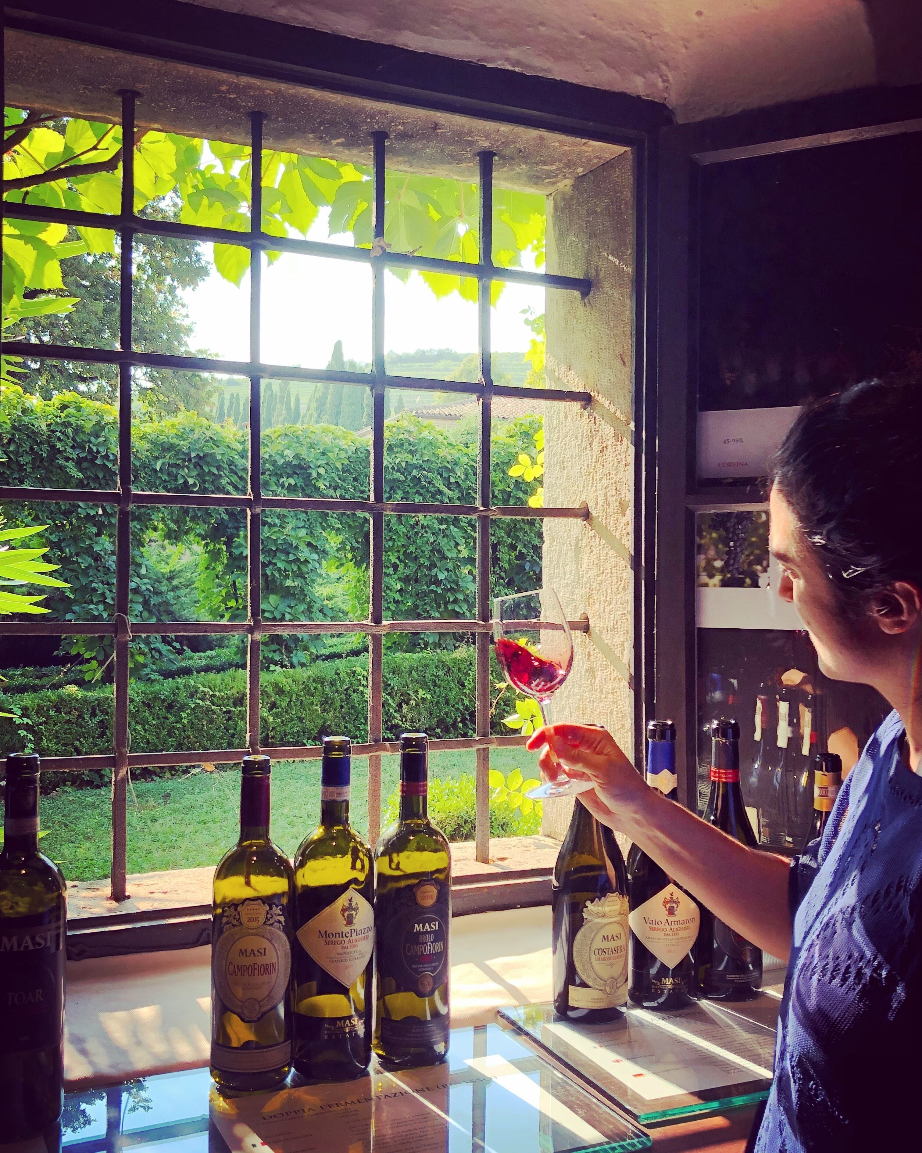 Una persona che tiene un bicchiere di vino vicino a una finestra con vista sul verde. Si vedono diverse bottiglie di vino sul bancone.