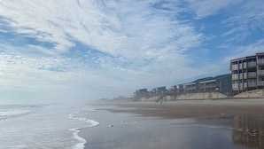 A beach with a row of multi-story buildings, some with balconies, and a clear sky with scattered clouds. - Oceanfront! Topsail Dunes Beachfront Condo with Pool and Balcony (North Topsail Beach)