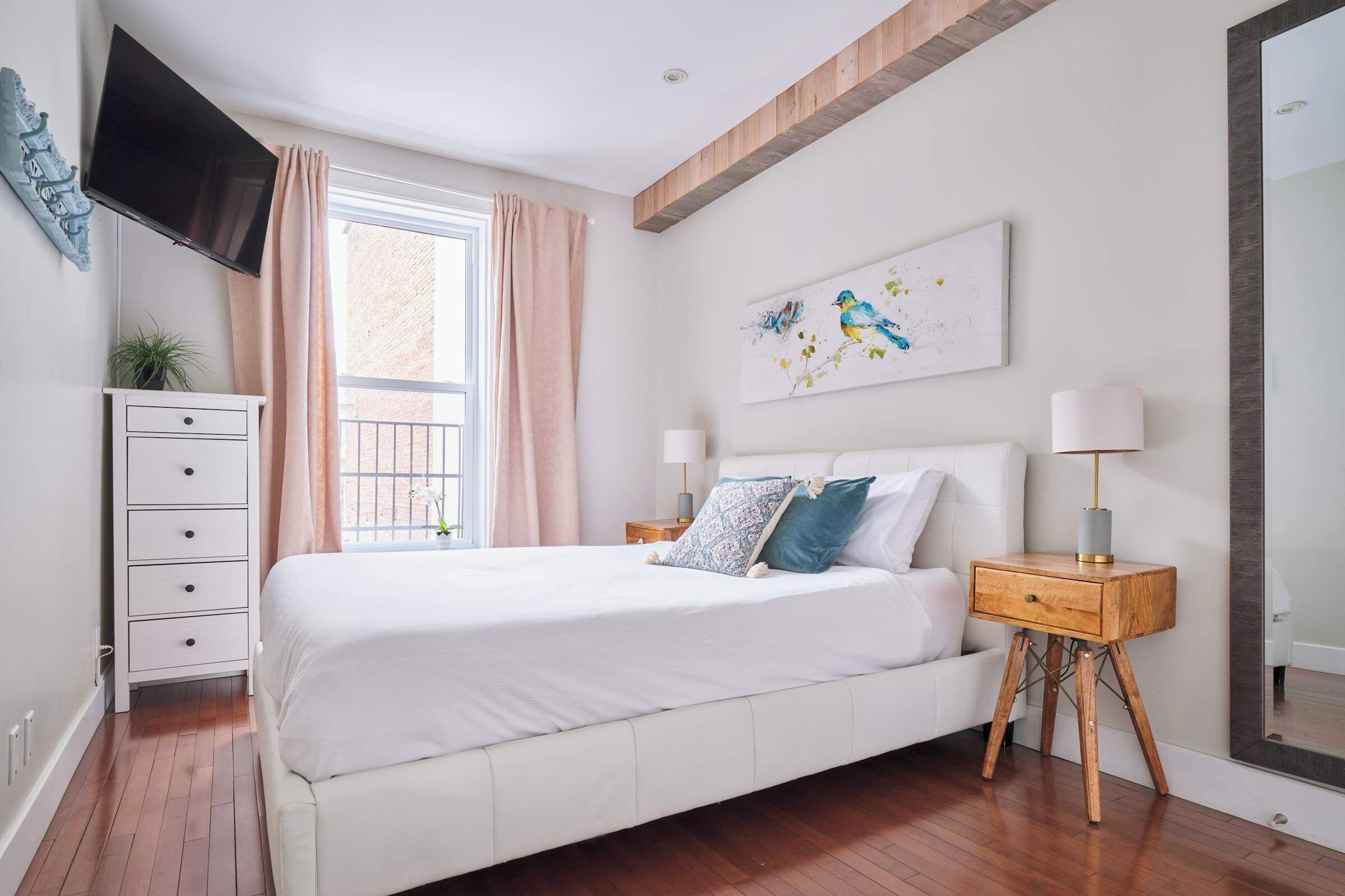 Un dormitorio con una cama grande, un televisor montado en la pared, una mesita de noche con una lámpara y un ventanal con cortinas.
