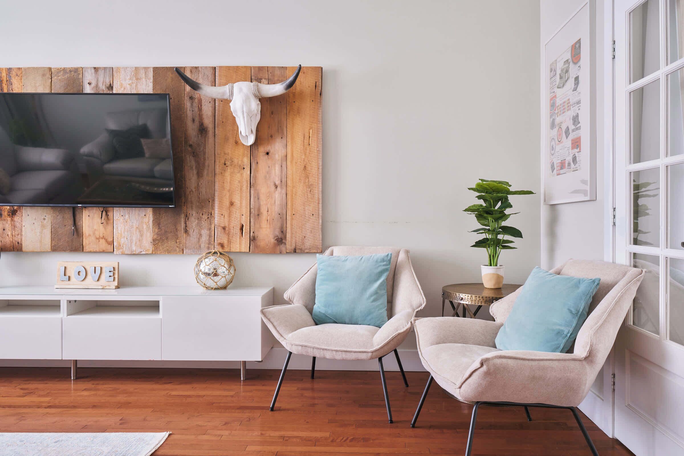 Ein modernes Wohnzimmer mit einer Holzvertäfelung, einem Wand-Fernseher, einer weißen Konsole, zwei beigen Sesseln mit blauen Kissen und einer Topfpflanze.