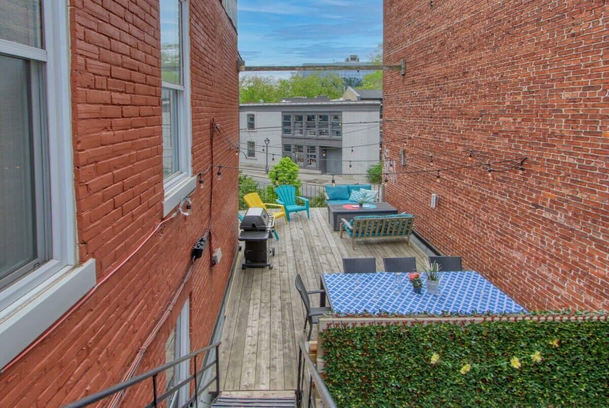 Un estrecho callejón con un muro de ladrillo a un lado y una terraza de madera al otro. Hay una mesa azul con diseño blanco, una silla verde y una silla amarilla.