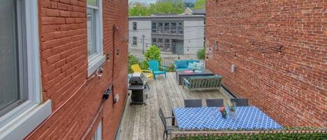 Un estrecho callejón con un muro de ladrillo a un lado y una terraza de madera al otro. Hay una mesa azul con diseño blanco, una silla verde y una silla amarilla.