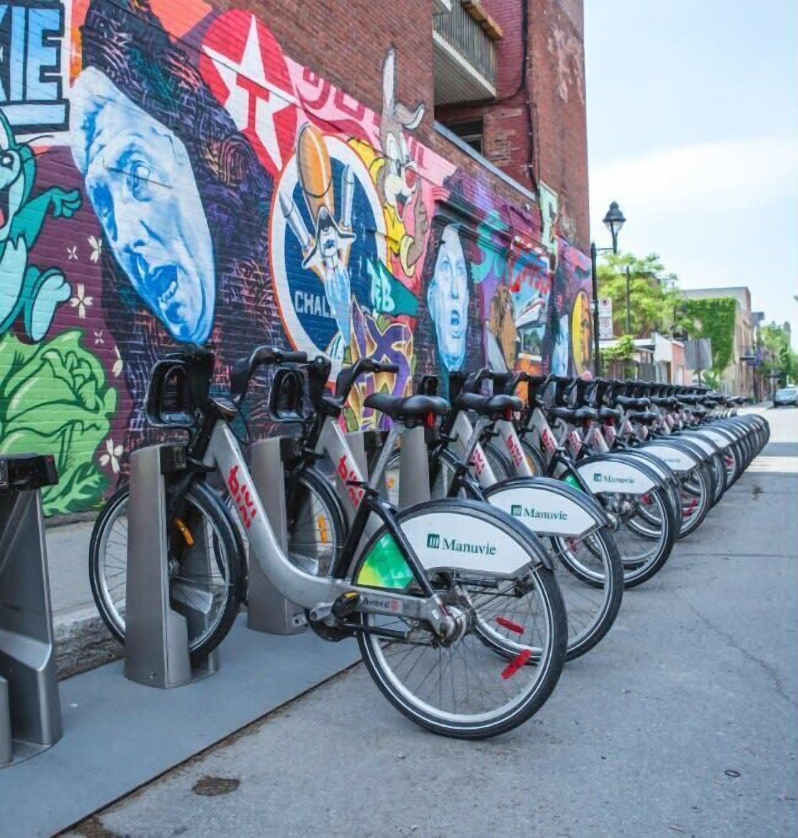 Une rangée de vélos électriques Manuvie est stationnée devant une fresque colorée sur un bâtiment en briques.