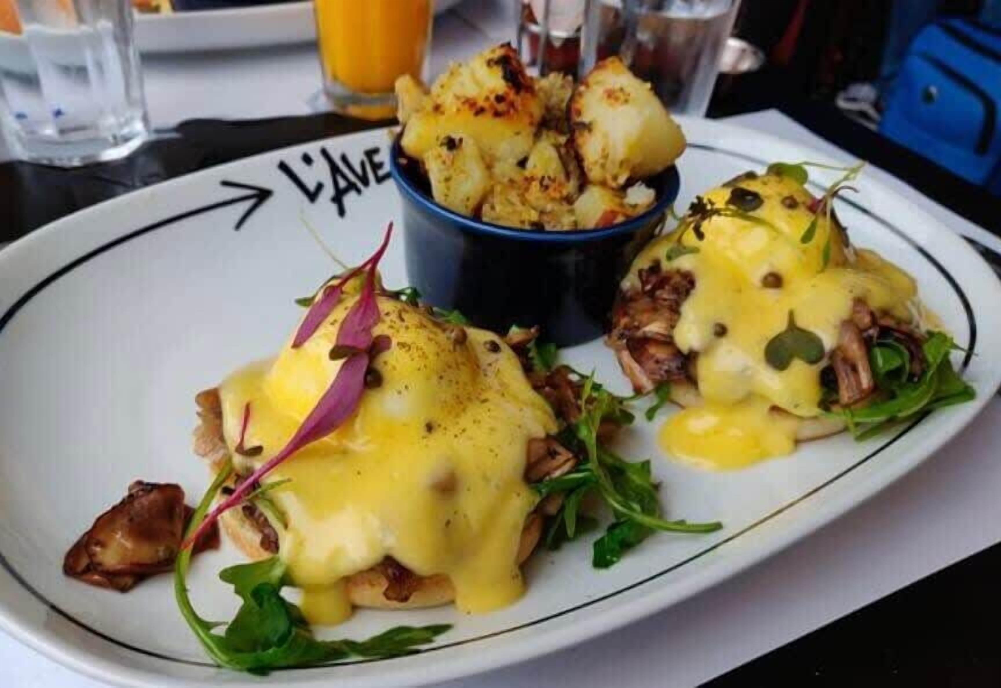 A plate of eggs benedict with hollandaise sauce, accompanied by a small bowl of roasted potatoes.