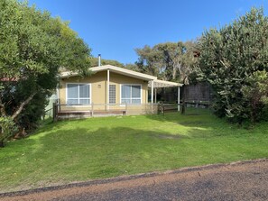 A single-story house with a covered patio, surrounded by a well-maintained lawn and dense trees. - Dog friendly close to beach/shops, trampoline, hot outside shower, NBN Wifi (Sandy Point)