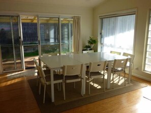 A dining area with a white table and chairs, large windows, and a view of greenery outside. - Dog friendly close to beach/shops, trampoline, hot outside shower, NBN Wifi (Sandy Point)