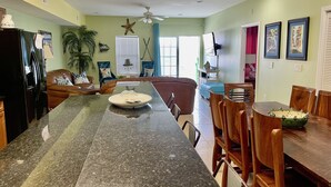 A kitchen with a granite countertop, a dining area with wooden chairs, and a living room with a sofa and a TV. - Bubbles Up. Ocean View Corner Unit in Coconut Grove. North Myrtle Beach, SC (North Myrtle Beach)