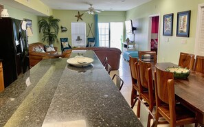 A kitchen with a granite countertop, a dining area with wooden chairs, and a living room with a sofa and a TV.
