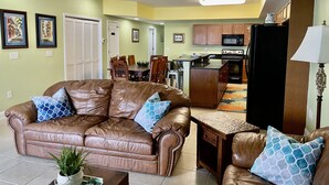 A living room with a brown leather sofa, a coffee table, and a dining area with wooden chairs and a table. - Bubbles Up. Ocean View Corner Unit in Coconut Grove. North Myrtle Beach, SC (North Myrtle Beach)