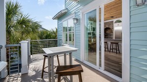 Une terrasse couverte avec une table et des chaises, une porte-fenêtre coulissante donnant sur un coin cuisine équipé d’un four à pizza, et une vue sur des palmiers.
