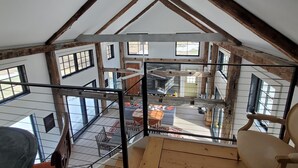 A high-ceilinged attic room with exposed wooden beams, a glass railing, and a view of the lower level.