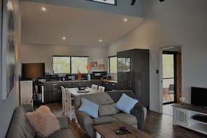 Ein modernes Wohnzimmer mit grauem Sofa, einem Couchtisch und einer TV-Kommode. Es gibt eine Küchenzeile mit Esstisch und Stühlen, und ein großes Fenster sorgt für natürliches Licht.