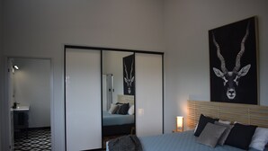 Une chambre à coucher avec un lit, un grand miroir, une œuvre d’art accrochée au mur et une table de chevet avec une lampe.