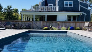 A rectangular swimming pool with clear water, surrounded by a wooden deck and a house with a balcony in the background.