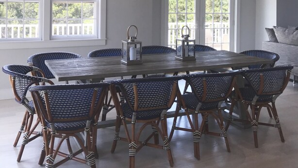 A dining area with a large wooden table surrounded by wicker chairs, two hanging lanterns, and large windows with a view of greenery.