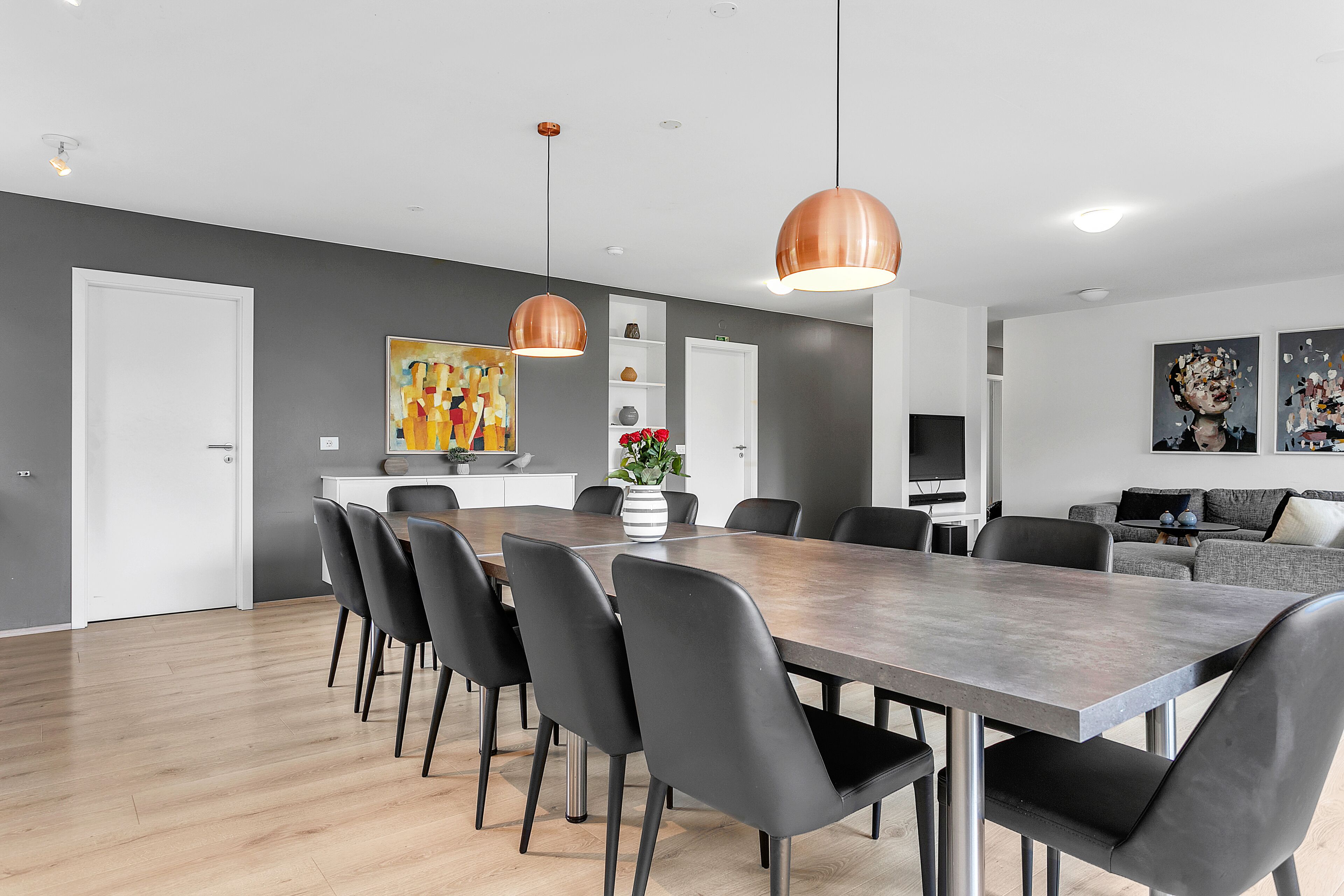 Uma sala de jantar moderna com uma mesa comprida, cadeiras pretas e luminárias pendentes em cobre.