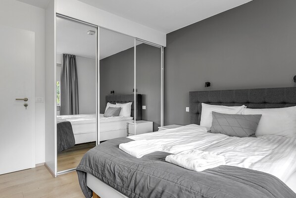 A modern bedroom with a large bed, a wardrobe with a mirror, and a bedside table.